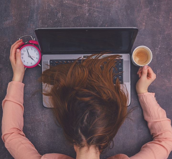 Frau legt Kopf auf den Laptop, in den Händen eine Uhr und eine Kaffeeetasse