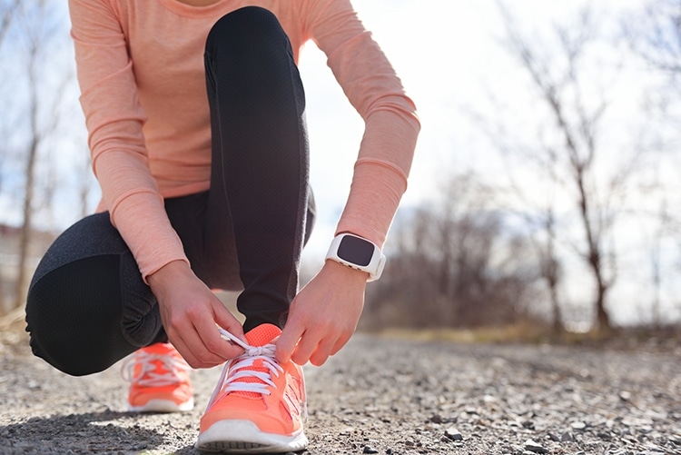 Running shoes and runner sports smartwatch. Female runner tying shoe laces on running trail using smart watch heart rate monitor.